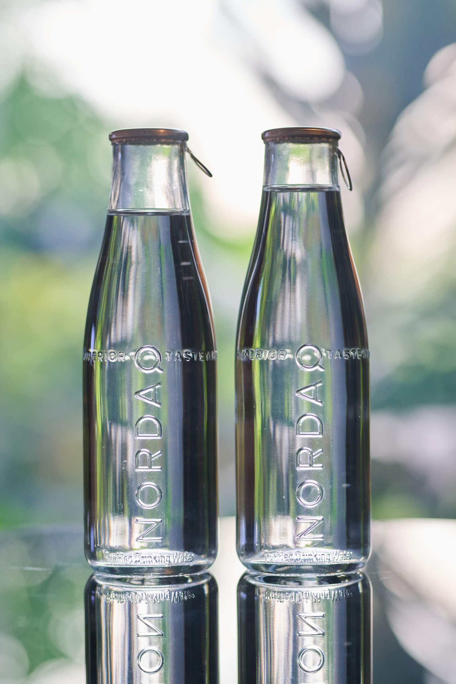 Two Nordaq glass bottles of water on a mirrored table, green garden light blurred behind them
