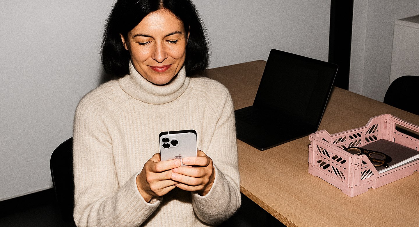 A woman in a cream turtleneck smiling at her phone at an office desk, a laptop and a pink crate of papers beside her
