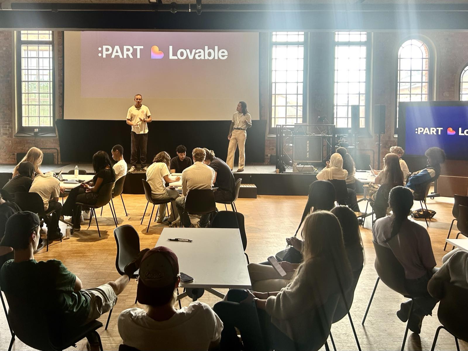 The AI workshop at Berghs: two facilitators on stage under the :PART and Lovable logos, teams at tables in front